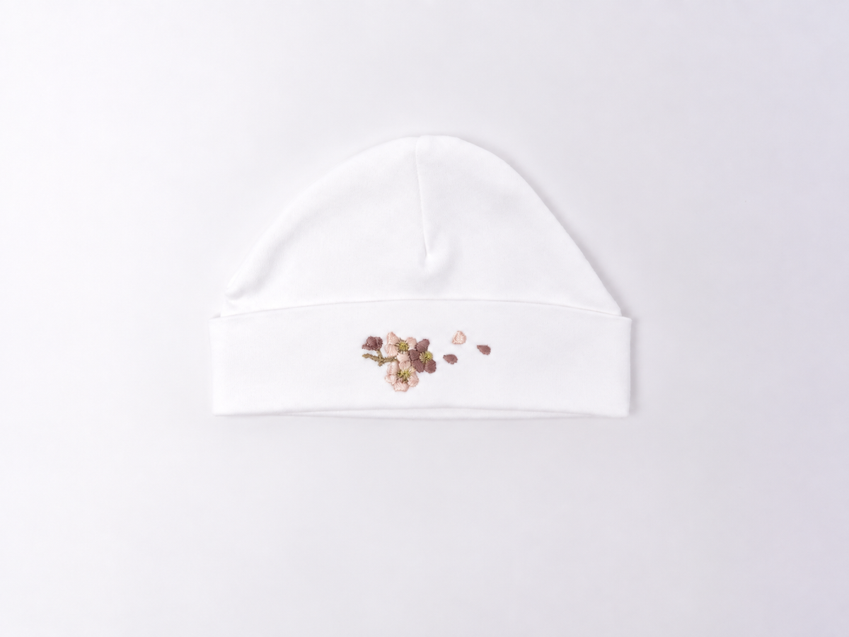 A white cotton baby beanie hat featuring a small floral and heart-shaped sequin embellishment on the front of the folded cuff, displayed on a clean white background.