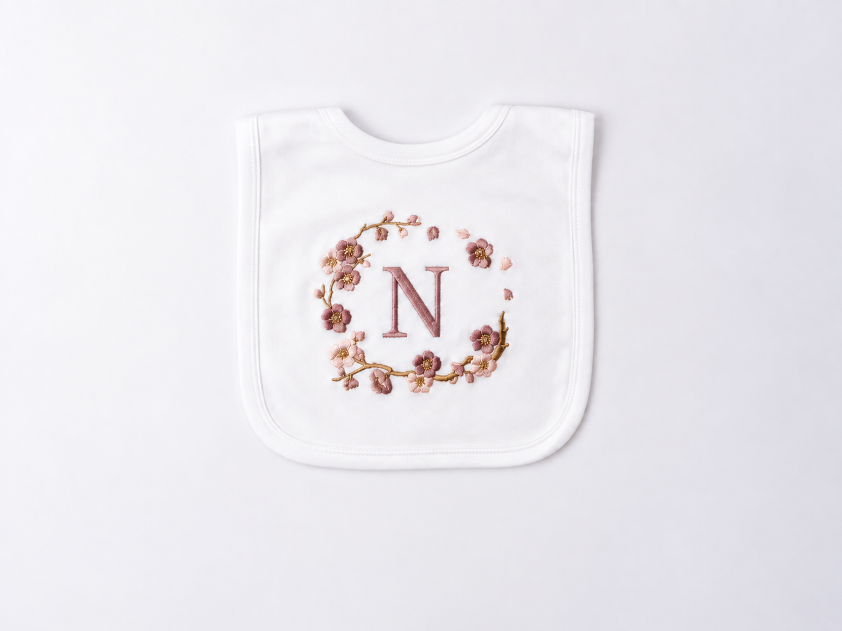 A white baby bib with a personalised design featuring a rose gold embroidered letter 'N' enclosed in a delicate floral blossom wreath, displayed on a clean white background.