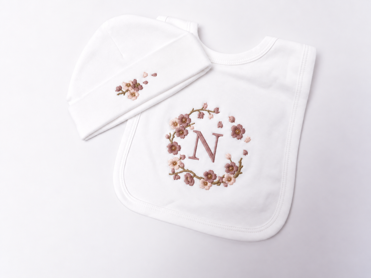 A white baby bib and matching beanie set with delicate floral blossom embroidery. The bib features a central floral wreath design surrounding a monogram letter 'N' in dusty rose, while the matching beanie has a smaller version of the floral embroidery on the front.