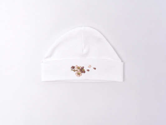 A white cotton baby beanie hat featuring a small floral and heart-shaped sequin embellishment on the front of the folded cuff, displayed on a clean white background.