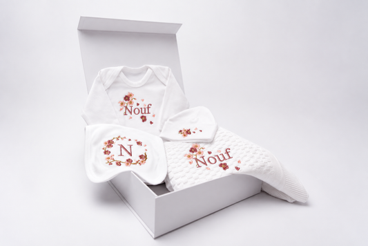 A white baby gift set including a onesie, a hat, a bib, and a blanket, all personalised with pink floral blossom embroidery and Nouf embroidered ('Nouf' and 'N'), presented in an open white box