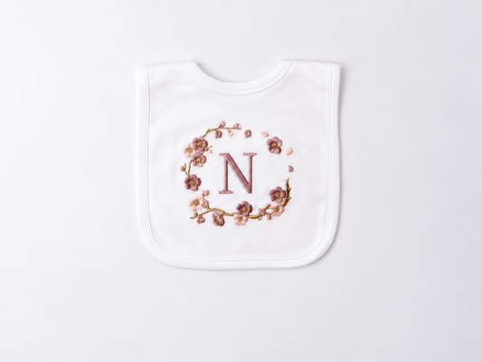 A white baby bib with a personalised design featuring a rose gold embroidered letter 'N' enclosed in a delicate floral blossom wreath, displayed on a clean white background.