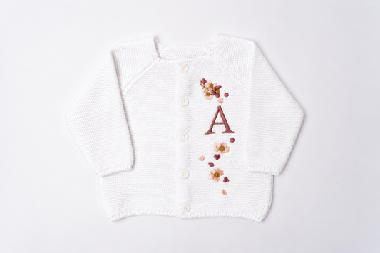 A white knit baby cardigan sweater with a button-up front, featuring a personalized embroidered letter 'A' and small floral accents in brown, pink, and yellow.