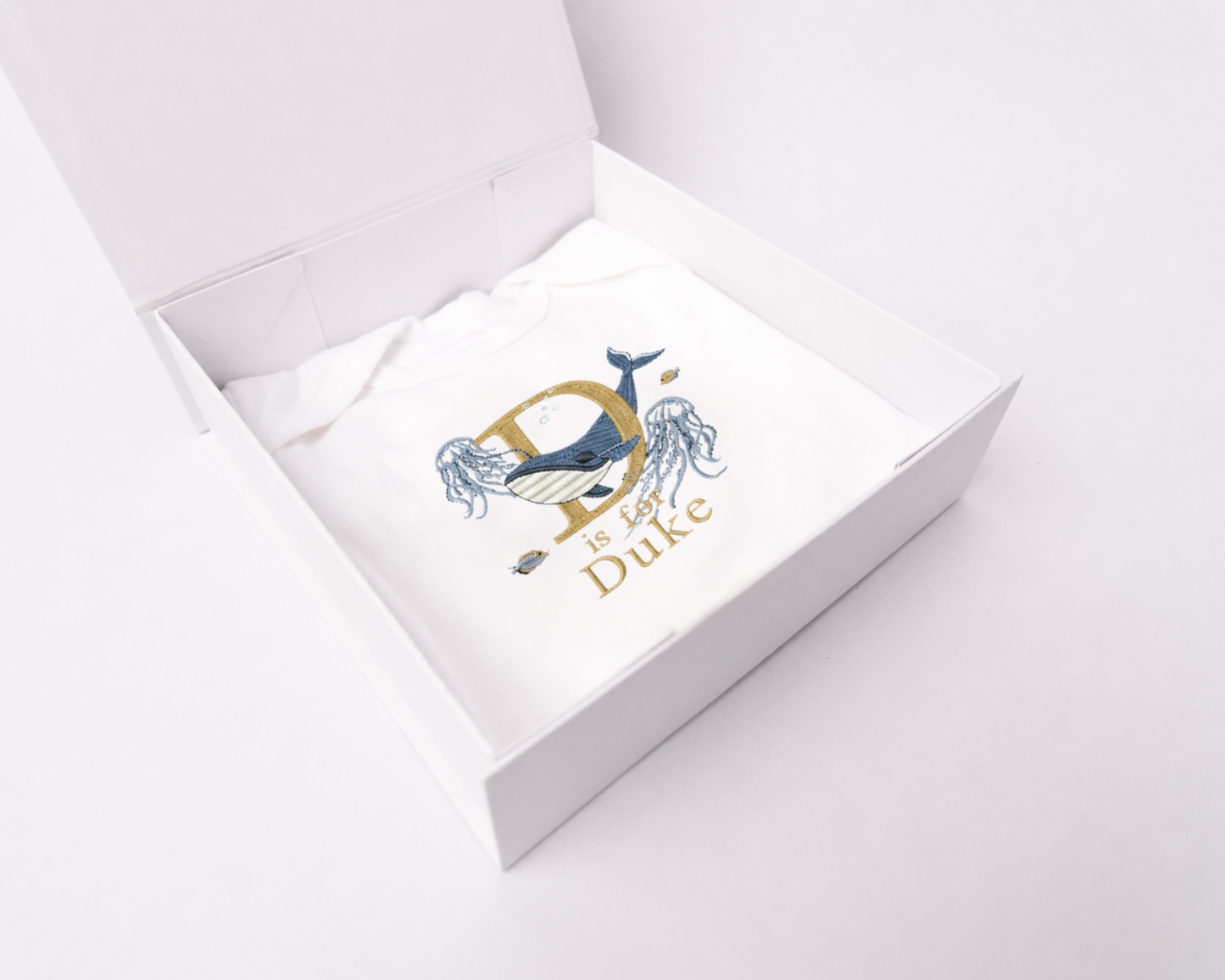 A white square gift box shown open from an elevated angle, revealing a folded white romper inside with an embroidered underwater world. The embroidery features a gold letter D, a blue and white whale, and the personalised name 'Duke'