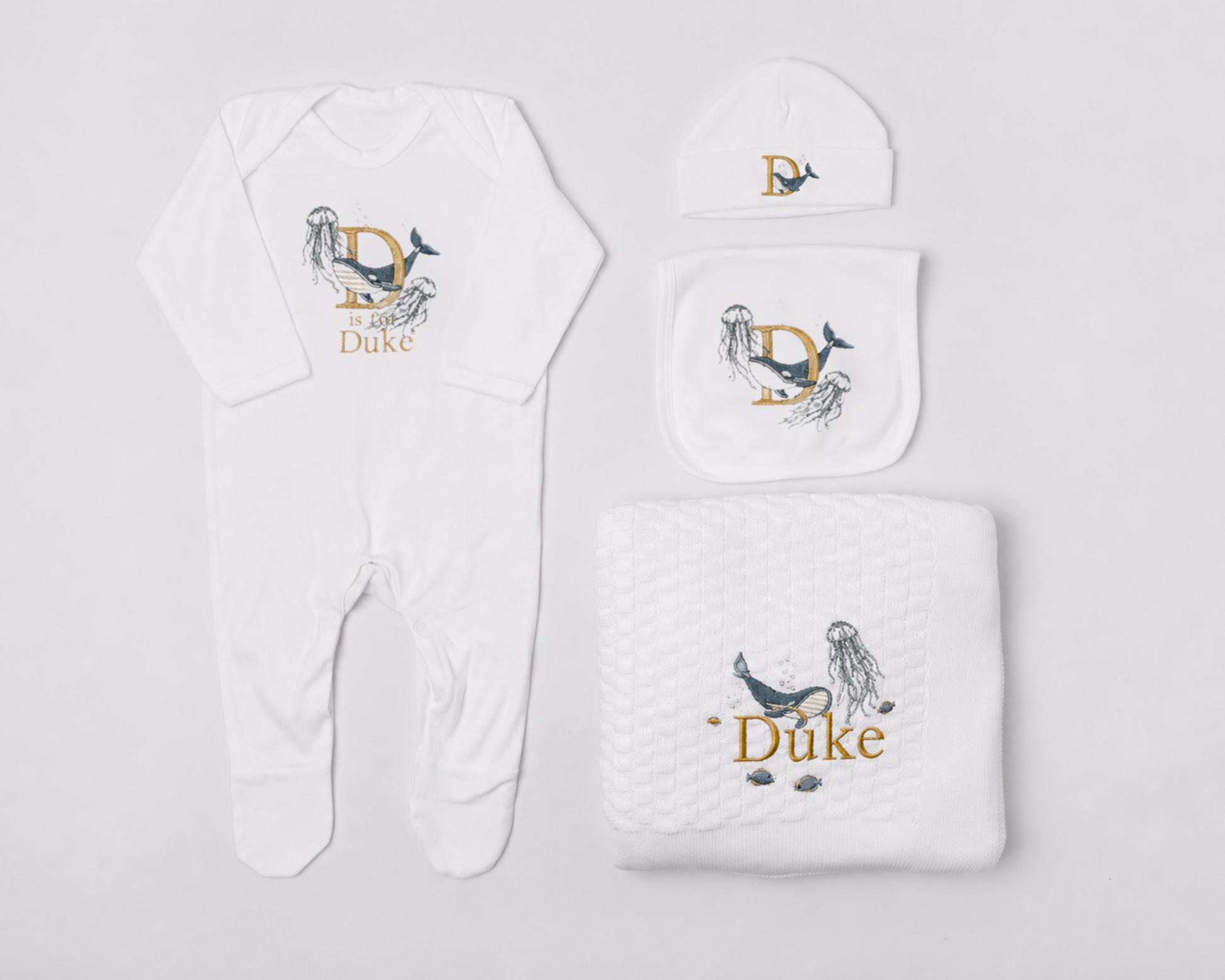 A white baby gift set including a footed romper, a beanie, a bib, and a blanket, all featuring a gold and blue underwater world embroidery, personalised for a baby named Duke
